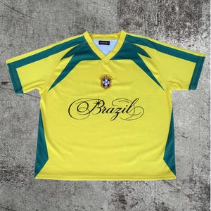May include: A yellow short-sleeved V-neck jersey with green accents on the shoulders and sides. The chest features a central crest with five stars and the word Brazil written in a black cursive script with a small star icon above the letter i.