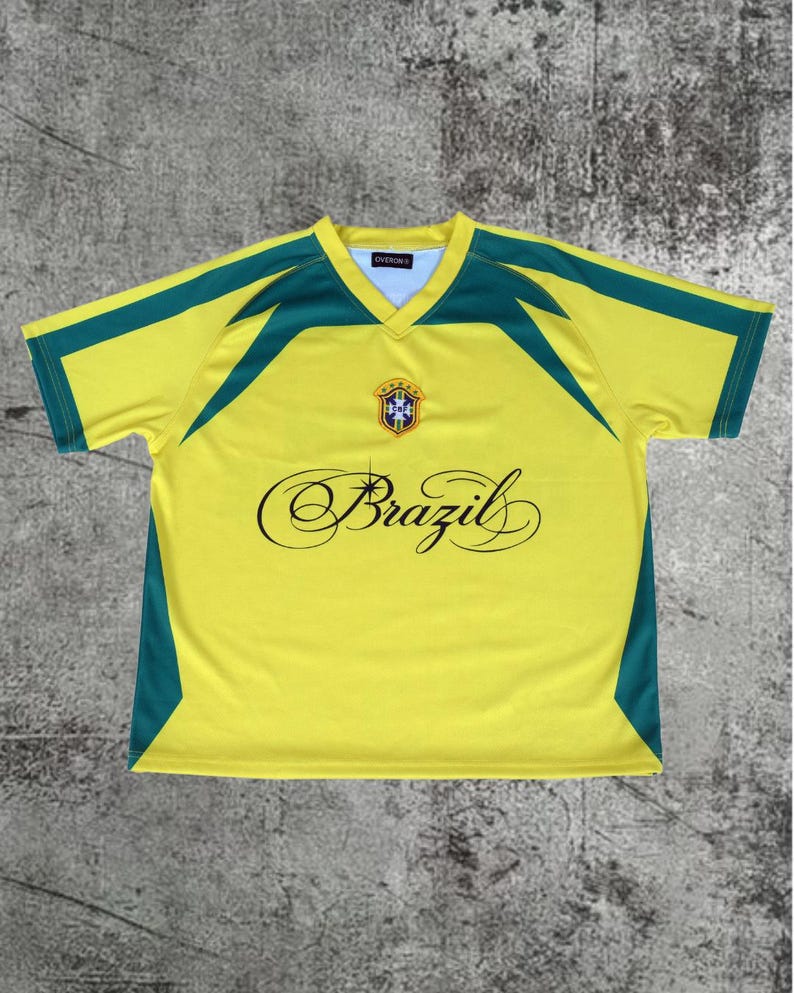 May include: A yellow short-sleeved V-neck jersey with green accents on the shoulders and sides. The chest features a central crest with five stars and the word Brazil written in a black cursive script with a small star icon above the letter i.