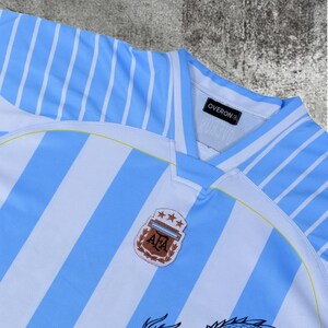 May include: A light blue and white striped sports jersey featuring a V-neck collar with an Overon brand tag inside. The chest displays an AFA crest with three stars above it, and a partial black dragon graphic is visible at the bottom edge.