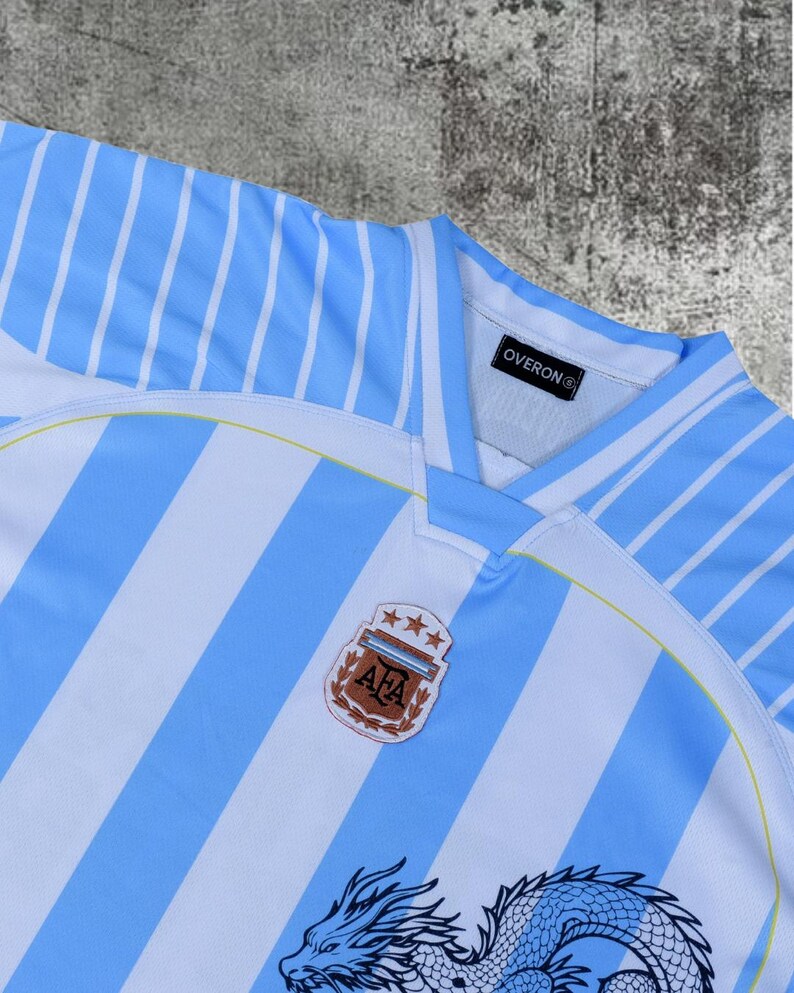 May include: A light blue and white striped sports jersey featuring a V-neck collar with an Overon brand tag inside. The chest displays an AFA crest with three stars above it, and a partial black dragon graphic is visible at the bottom edge.