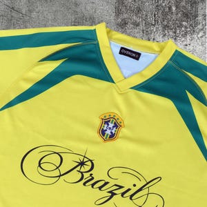 May include: A yellow jersey with dark green accents on the shoulders and a V-neck collar. The chest features a CBF crest with five stars and the word Brazil in a black cursive script. The interior neck label displays the brand name OVERON.