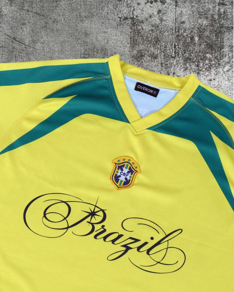 May include: A yellow jersey with dark green accents on the shoulders and a V-neck collar. The chest features a CBF crest with five stars and the word Brazil in a black cursive script. The interior neck label displays the brand name OVERON.