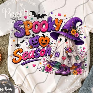 Op de afbeelding: Een wit shirt met lange mouwen en een kleurrijke afbeelding van een geest met een paarse heksenhoed en laarzen, omringd door bloemen. De tekst Spooky Season staat in paarse en oranje letters op het shirt afgebeeld.
