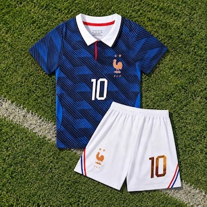 Könnte beinhalten: Ein blaues Fußballtrikot mit dunkelblauem Muster, weißem Kragen, der weißen Nummer 10 und einem goldenen Hahn-Emblem. Dazu eine weiße Fußballhose mit der goldenen Nummer 10, einem goldenen Hahn-Emblem sowie roten und blauen Seitenstreifen.