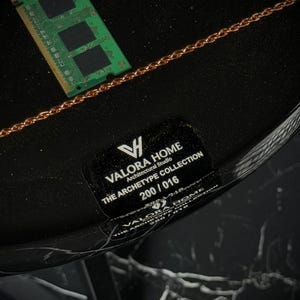 May include: A black circular surface featuring a green RAM stick and a copper-colored chain. A black label displays the text: VALORA HOME, Architectural Studio, THE ARCHETYPE COLLECTION, 200 / 016. The base has a black and white marble pattern.