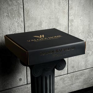 May include: A square black cardboard box with gold lettering. The top features the logo and text VALORA HOME Architectural Studio. The front side displays the text HANDCRAFTED IN ISTANBUL. The box is resting on a black fluted column pedestal.