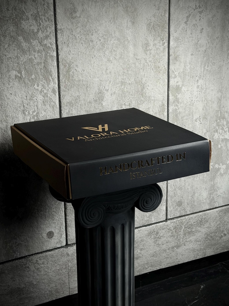 May include: A square black cardboard box with gold lettering. The top features the logo and text VALORA HOME Architectural Studio. The front side displays the text HANDCRAFTED IN ISTANBUL. The box is resting on a black fluted column pedestal.
