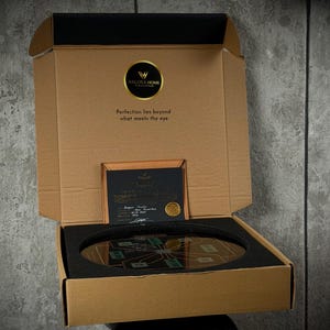 May include: A brown cardboard box with an open lid containing a circular object with a circuit board design, protected by black foam inserts. The lid features the Valora Home logo and the text, Perfection lies beyond what meets the eye.