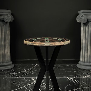 May include: A round side table with a black top featuring a geometric pattern of green and copper-colored accents. The table has a black metal base with four intersecting legs that form an X-shape. The edge of the tabletop shows layered materials.