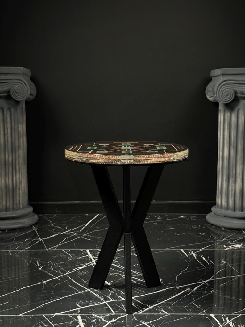 May include: A round side table with a black top featuring a geometric pattern of green and copper-colored accents. The table has a black metal base with four intersecting legs that form an X-shape. The edge of the tabletop shows layered materials.