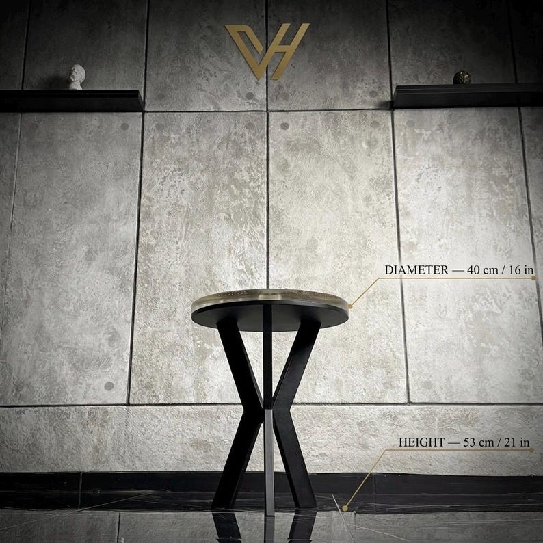 May include: A round side table with a dark circular top and a black metal base featuring crossed legs. The table has a diameter of 16 inches and a height of 21 inches. The design includes a flat surface supported by a sturdy, angular frame structure.