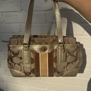 Puede incluir: Un bolso de mano Coach con patrón de lona monograma marrón, adornado con ribetes y asas de cuero dorado metalizado. La parte frontal presenta una franja vertical central en marrón, crema y beige con un cierre giratorio de metal dorado.