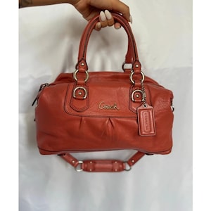 May include: A coral-colored leather handbag featuring two top handles, silver-tone metal hardware, and a detachable leather luggage tag. The front displays the Coach logo in metallic lettering, with a zippered closure and a shoulder strap.