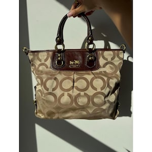Könnte beinhalten: Eine beige-braune Tragetasche mit einem sich wiederholenden kreisförmigen Logomuster auf dem Stoffkörper. Die Tasche hat dunkelbraune Lederbesätze, zwei Tragegriffe mit goldfarbenen Metallbeschlägen und ein goldenes Coach-Logo auf der Vorderseite.