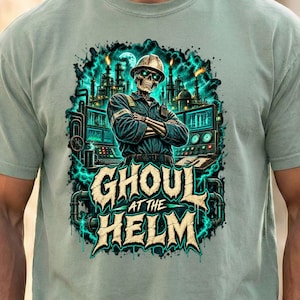 May include: A sage green short-sleeve t-shirt featuring a graphic print of a skeleton wearing a hard hat and uniform with arms crossed. The design includes industrial machinery, glowing teal energy, and the text GHOUL AT THE HELM in bold letters.
