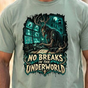May include: A sage green short-sleeve t-shirt featuring a graphic print of a deer-headed figure sitting at a desk with multiple computer monitors. The text reads NO BREAKS IN THE UNDERWORLD. The design includes dark forest elements and a skull.