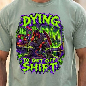 May include: A sage green short-sleeve t-shirt featuring a graphic print of a red-skinned demon sitting at a control panel in a factory. The text DYING TO GET OFF SHIFT is printed in bright green, dripping letters above and below the illustration.