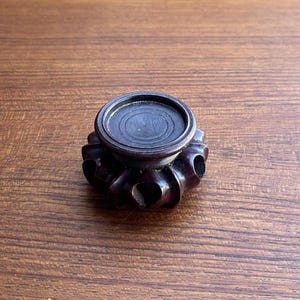 Puede incluir: Un pequeño soporte de exhibición de madera marrón oscuro con una parte superior circular y una base festoneada similar a pétalos. El soporte presenta una plataforma central lisa y plana rodeada de protuberancias redondeadas talladas.