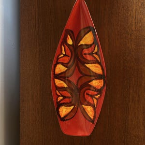 Peut inclure: Un plat en céramique en forme de feuille allongée avec une base de couleur orange vif. Le centre du plat est orné d'un motif abstrait symétrique dans des tons de brun foncé et de jaune doré, créant un contraste visuel audacieux et saisissant.