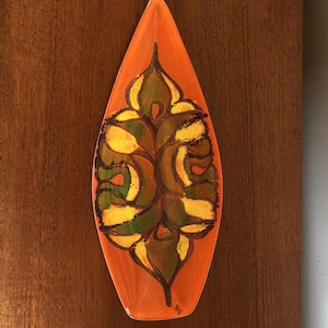 Könnte beinhalten: Eine längliche, blattförmige Keramikschale mit einer leuchtend orangefarbenen Grundfarbe. Die Mitte der Schale ist mit einem symmetrischen, abstrakten Muster in Dunkelgrün, Senfgelb und Schwarz verziert, das ein kräftiges, organisches Design bildet.
