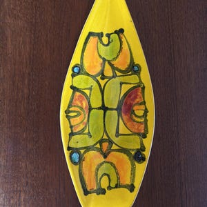 Könnte beinhalten: Eine spitz zulaufende, ovale Keramikschale mit leuchtend gelber Glasur. In der Mitte befindet sich ein symmetrisches, abstraktes Grafikdesign in Limettengrün, Orange und Schwarz, akzentuiert durch kleine, kreisförmige türkise und dunkelblaue Punkte.