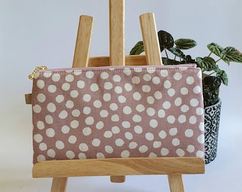 May include: A small pink zippered pouch with a white polka dot pattern. The pouch is displayed on a wooden easel.
