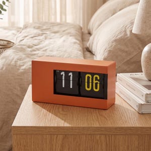 May include: A rectangular orange flip clock with a black display panel showing the time 11:06 AM. The clock features white numerals for the hour and yellow numerals for the minutes, set within a solid orange casing with a smooth matte finish.