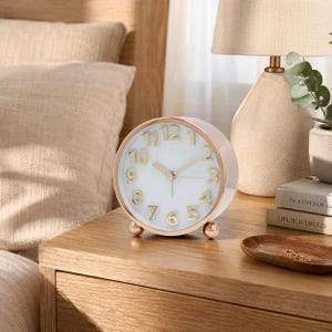 May include: A round analog alarm clock with a white face, gold-toned numbers, and gold-toned hands. The clock features a metallic rose gold-colored frame and rests on two small, spherical gold-toned feet. The design is simple and circular.