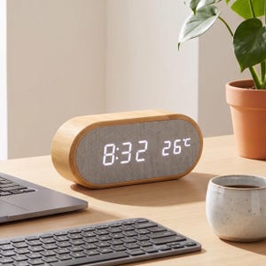 May include: An oval-shaped digital alarm clock with a light wood grain frame and a grey fabric front panel. The clock features a bright white LED display showing the time as 8:32 and the temperature as 26 degrees Celsius on its front face.