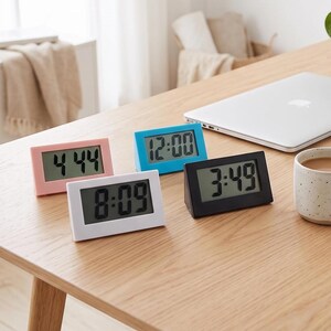 May include: Four rectangular digital clocks with slanted frames in pink, blue, white, and black. Each clock features a clear LCD screen displaying black numerical time digits. The clocks are arranged on a wooden surface, showing times 4:44, 12:00, 8:09, and 3:49.