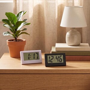 May include: Two rectangular digital clocks with LCD screens, one in a white frame displaying 1:17 and one in a black frame displaying 12:46. The clocks are positioned on a wooden surface next to a potted green plant and a table lamp.