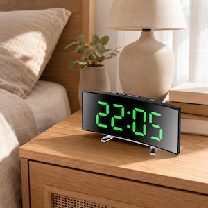 Può includere: Una sveglia digitale curva con una superficie a specchio nera riflettente e numeri LED verde brillante che mostrano le 22:05. L'orologio presenta un design rettangolare elegante con piccoli piedini in plastica trasparente per la stabilità.