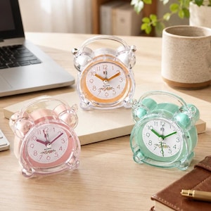 May include: Three small twin-bell alarm clocks with transparent plastic casings. One clock has a pink face with pink hands, one has an orange face with orange hands, and one has a white face with green hands. Each clock features the text Quartz.