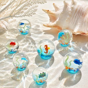 May include: A collection of seven clear glass spheres, each containing a miniature aquatic scene with colorful glass sea creatures including a seahorse, crab, jellyfish, and fish, set against green glass seaweed and small pebbles.