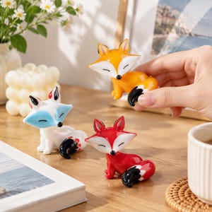 May include: Three small fox figurines made of smooth, glossy material. One is orange with a white face, one is white with light blue accents, and one is bright red. Each fox has closed eyes, a black nose, and a curled tail with black tips.