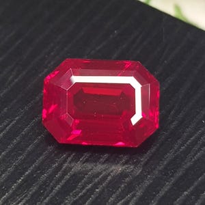 May include: A vibrant, deep red gemstone cut in a rectangular emerald shape with faceted surfaces that reflect light. The stone displays a rich, saturated crimson hue throughout its structure, showcasing clear geometric lines and sharp, polished edges.