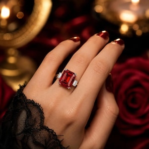 Puede incluir: Un anillo con una gran piedra preciosa roja rectangular de corte esmeralda montada en una banda de metal plateado. La piedra roja central está flanqueada a cada lado por una piedra transparente más pequeña de corte baguette rectangular.