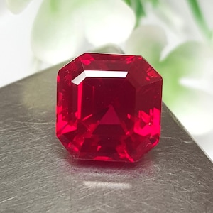 Könnte beinhalten: Ein leuchtend roter Edelstein im quadratischen Smaragdschliff mit scharfen, klaren Facetten, die das Licht reflektieren. Der Stein zeigt einen tiefen, gesättigten karmesinroten Farbton in seinem transparenten Körper und eine präzise Form.