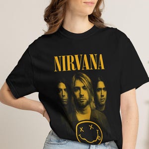 May include: A black short-sleeved t-shirt featuring a yellow graphic print of the band Nirvana. The design includes the word NIRVANA in a bold yellow font above a portrait of the three band members and a yellow smiley face icon with X eyes.