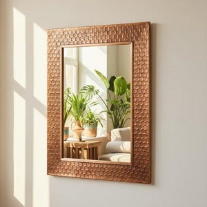 May include: A rectangular wall mirror featuring a metallic copper-toned frame with a textured, repeating honeycomb pattern. The reflective glass surface displays a clear view of indoor potted plants and furniture positioned near a bright window.