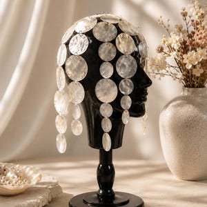 May include: A headpiece constructed from numerous translucent, circular mother-of-pearl discs linked together with small silver-toned metal rings. The discs vary in size and are arranged in cascading vertical strands that drape over a black mannequin head.