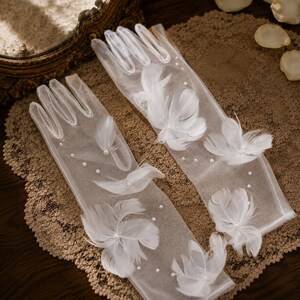 May include: A pair of sheer white tulle gloves featuring delicate white feather accents and small white faux pearls scattered across the fabric. The gloves extend to the wrist and are crafted from a lightweight, translucent material with finished edges.