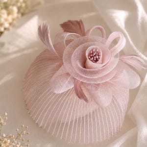 May include: A pale pink fascinator hat featuring a structured, pleated mesh base and a large, layered mesh rose embellishment at the center. The floral design is accented with small, matching pink beads in the middle and delicate, thin feathers.