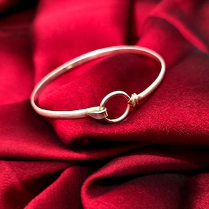 May include: A silver-toned bangle bracelet featuring a smooth, rounded band that terminates in a circular loop closure. The metal has a matte finish, and the design is characterized by a simple, continuous form that connects at the front with a small ring.