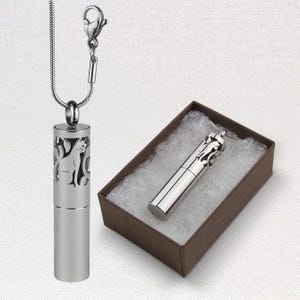May include: A silver-toned cylindrical pendant necklace featuring a cutout cat silhouette design. The pendant is attached to a snake chain with a lobster clasp. A matching pendant is displayed inside a brown rectangular box with white padding.