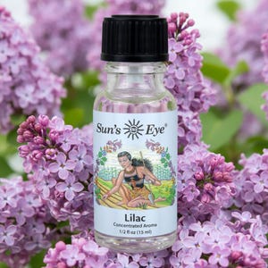 May include: A clear glass bottle with a black cap containing Sun's Eye Lilac concentrated aroma. The label features an illustration of a person in a field and states 1/2 fl oz (15 ml). The bottle is positioned in front of purple lilac flowers.