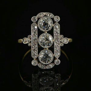May include: A gold ring featuring a vertical rectangular setting adorned with three prominent round-cut diamonds arranged in a column, surrounded by smaller pavé-set diamonds within a detailed metal frame that extends to the sides of the band.