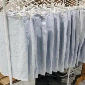 Puede incluir: Una fila de faldas de color azul claro con un sutil patrón floral tonal, colgadas en perchas de plástico blanco en un perchero de metal. Cada falda presenta una silueta recta y un acabado de tela texturizada, dispuestas en línea.