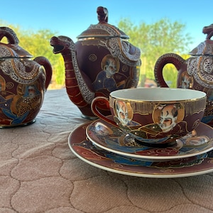 May include: A tea set featuring two teapots and a teacup with a saucer, all in a deep maroon color. Each piece is decorated with gold-trimmed illustrations of figures and dragon motifs. The teapots have lids topped with dragon-shaped finials.