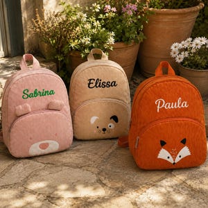 May include: Three corduroy backpacks featuring animal faces and embroidered names. The pink backpack displays the name Sabrina, the beige backpack shows the name Elissa with a dog face, and the orange backpack features the name Paula with a fox face.